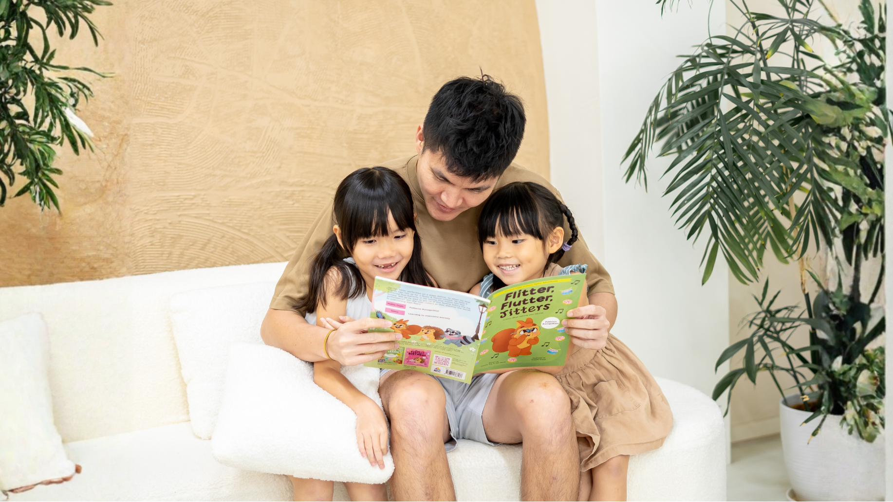 Man reading a book to two children on a white couch with plants in the background